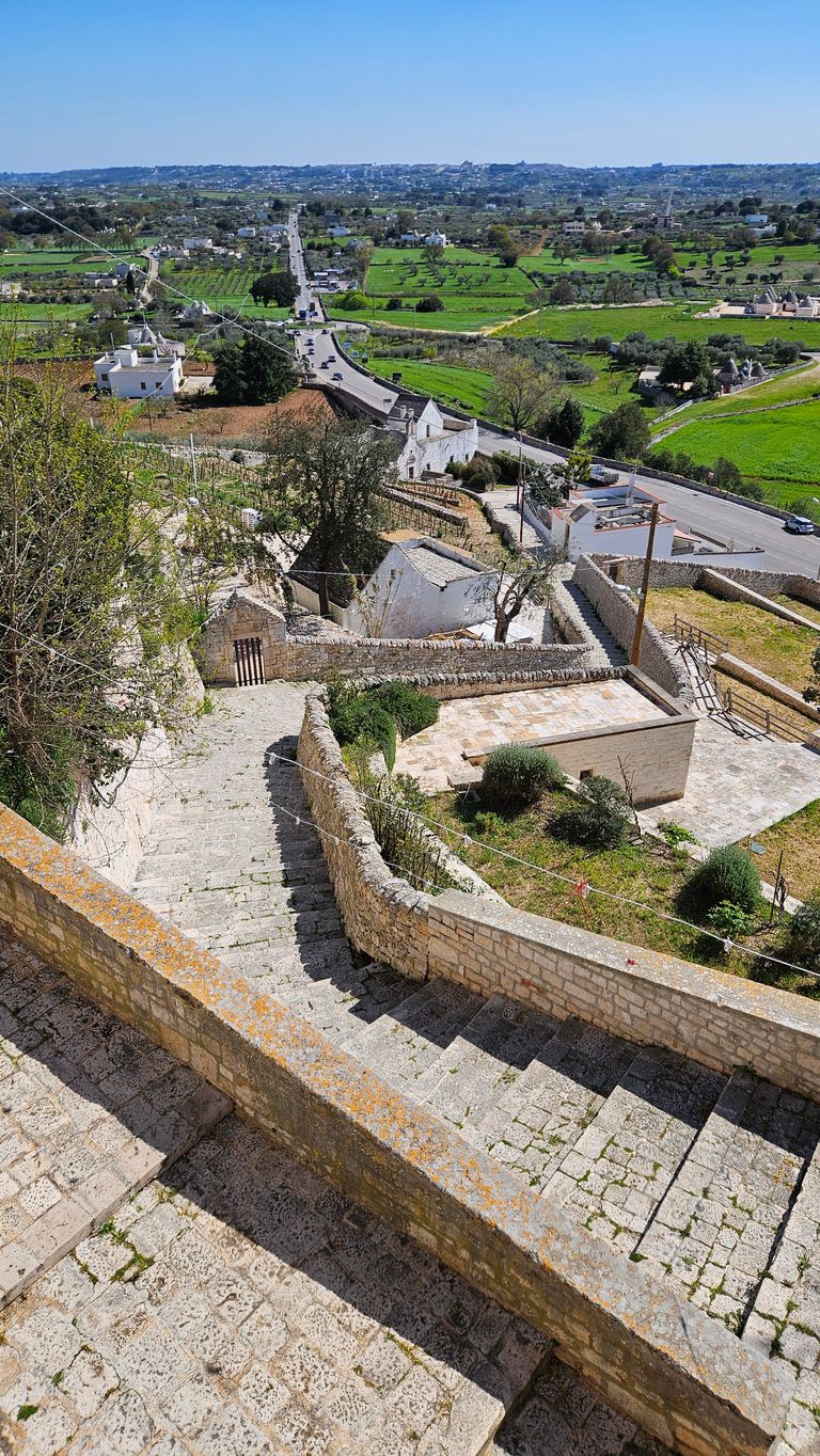 A stone staircase with low railing leading down to several white houses in a rural area with green fields and a road in the background.