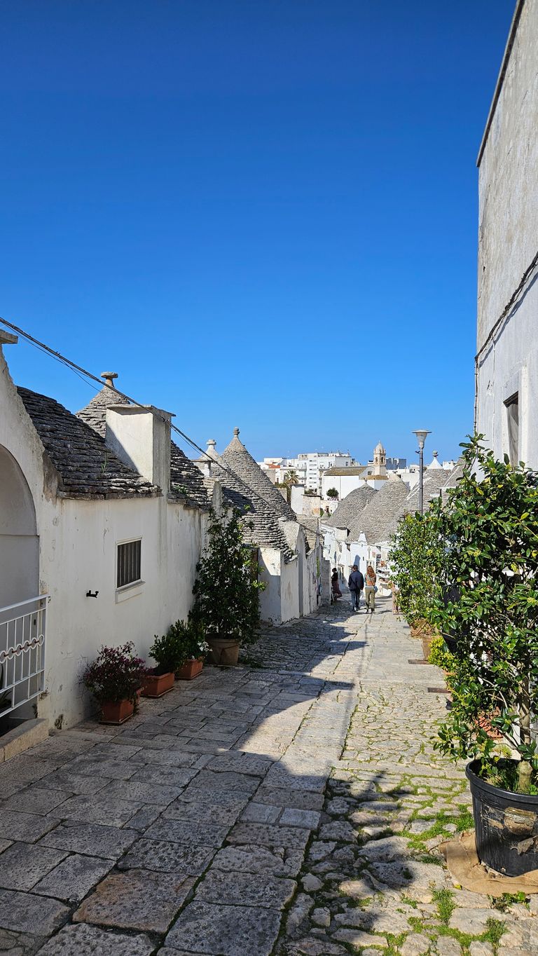 A paved street with traditional white houses and conical stone roofs under a clear blue sky, with some people walking at the end of the path.