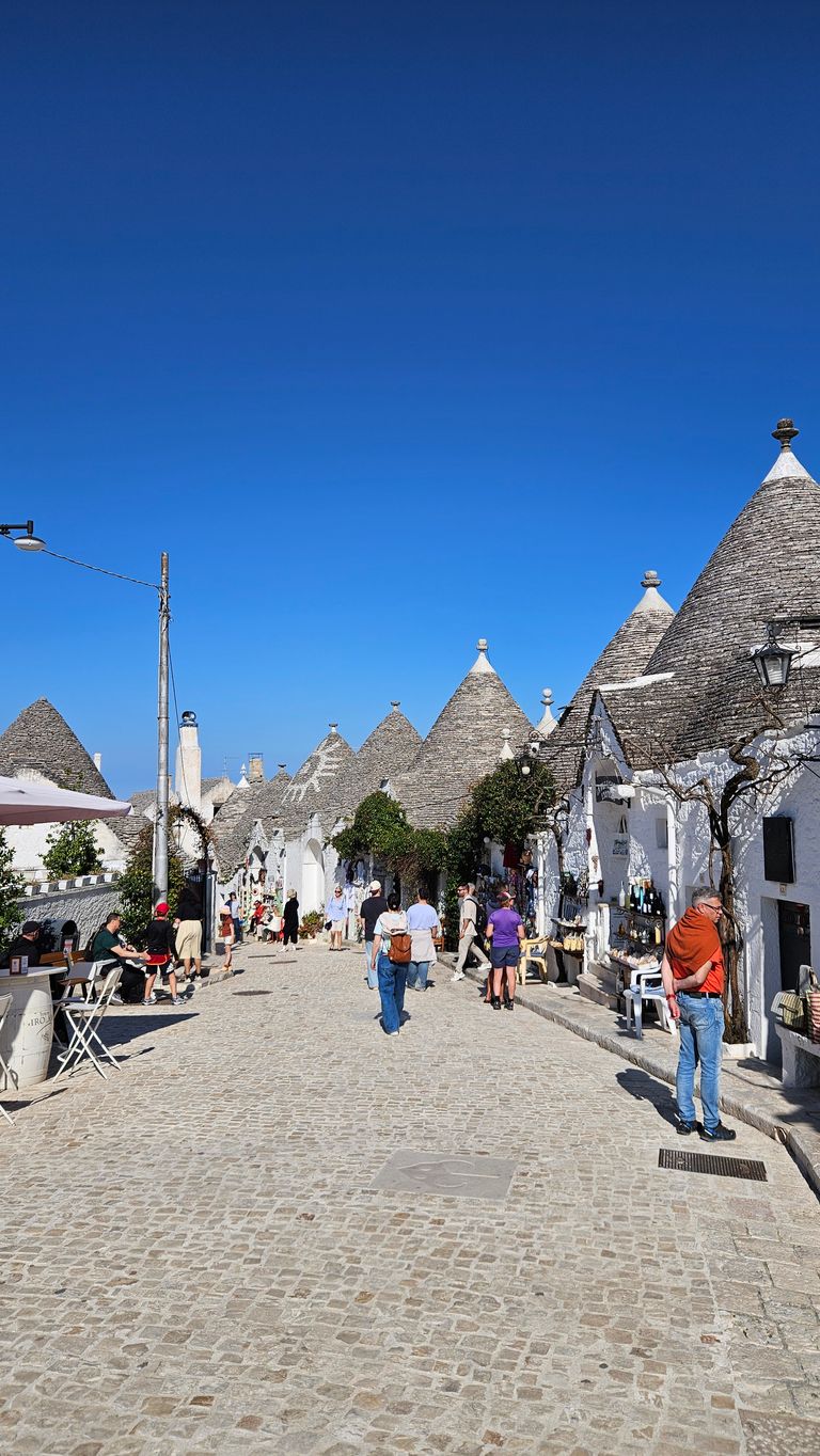 People walking on a cobblestone street lined with traditional white trullo houses with conical roofs under a clear blue sky.