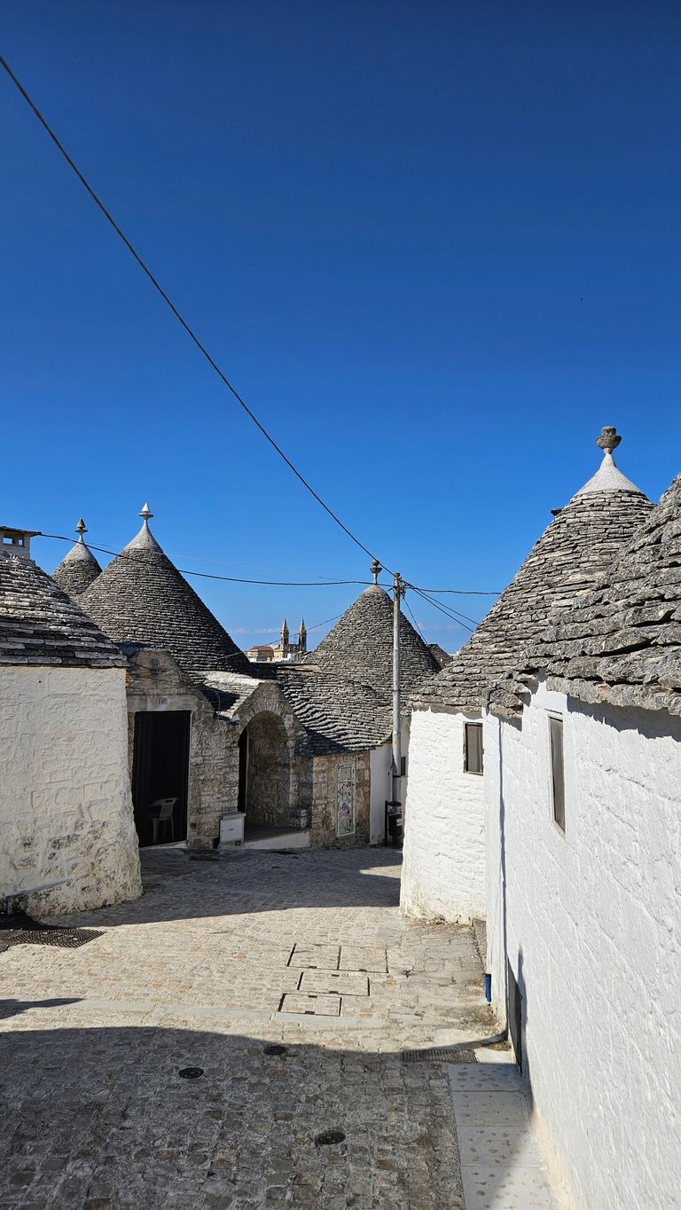 A cobblestone street lined with traditional stone houses featuring conical roofs under a clear blue sky.