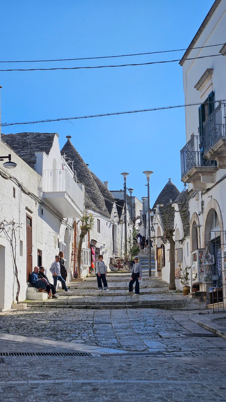 Cobblestone street with white houses and distinctive Trulli roofs under a clear blue sky, with several people standing and walking on the street.