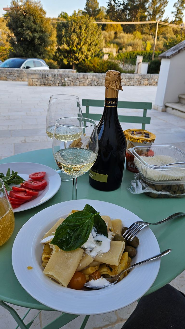 Table with a plate of pasta topped with fresh basil, two glasses of white wine, a bottle of sparkling wine, sliced tomatoes, and other food items outdoors in front of a stone wall and trees.
