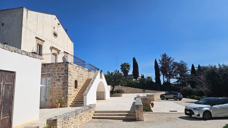 A sunny courtyard with a stone staircase leading to an elevated section of a white building with stone walls. Two cars are parked on a paved lot, surrounded by trees under a clear blue sky.