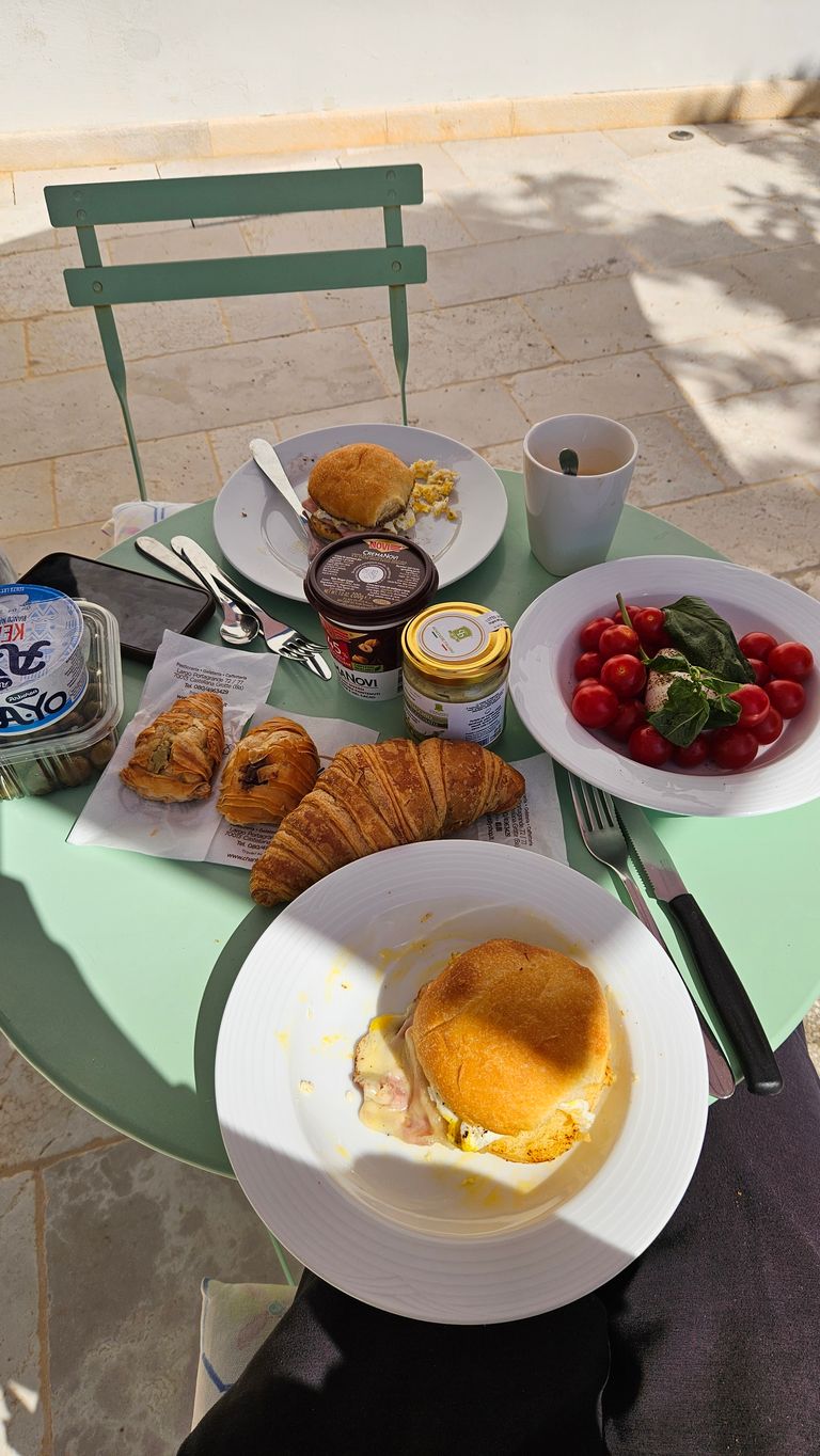 A green outdoor table with breakfast items including sandwiches on white plates, a croissant, small pastries, a bowl of cherry tomatoes with green leaves, a cup, and various small jars and containers.