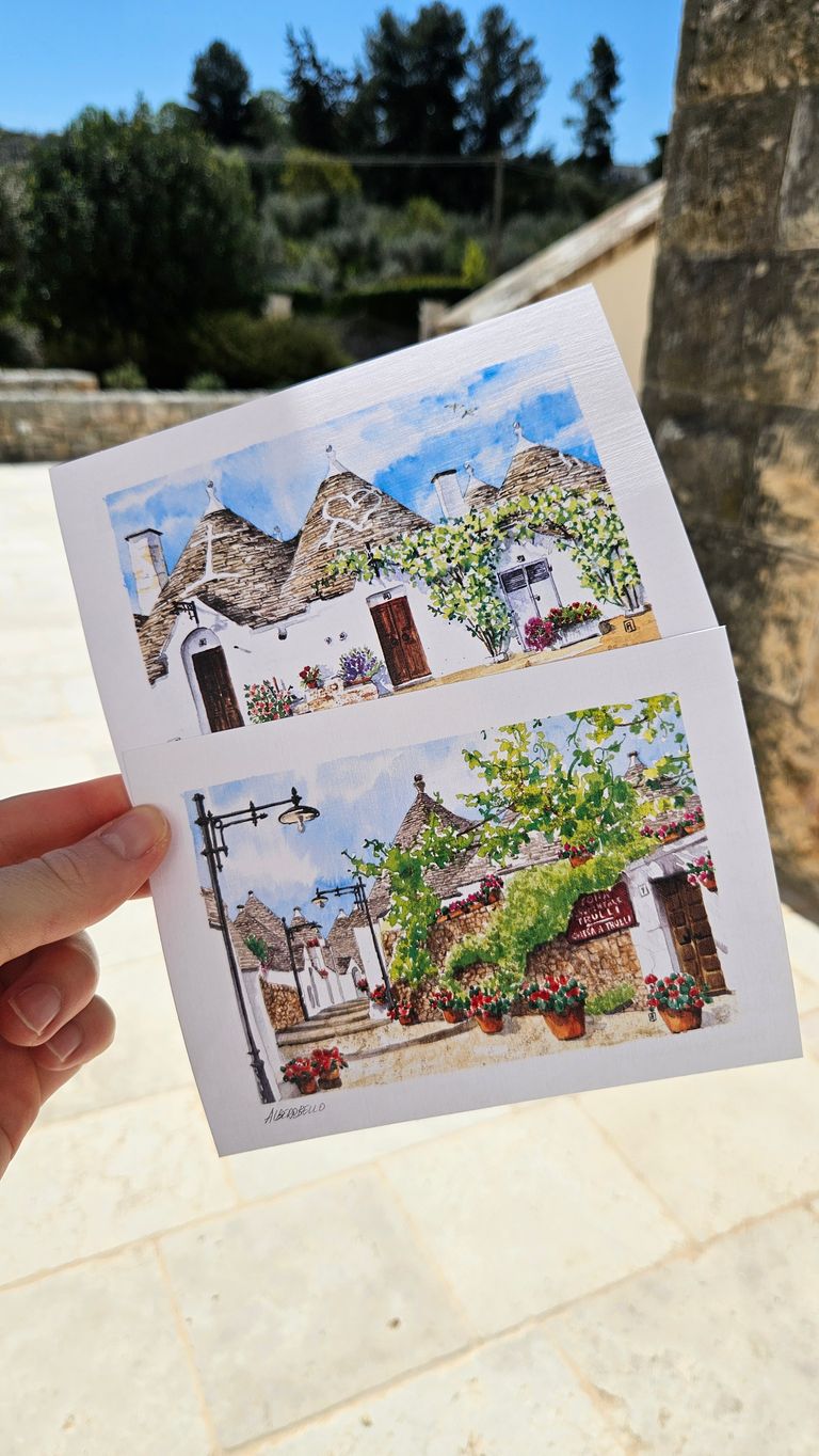 Hand holding two watercolor paintings of traditional Trulli houses with conical roofs, towers, and planted flowers in a sunny outdoor setting.