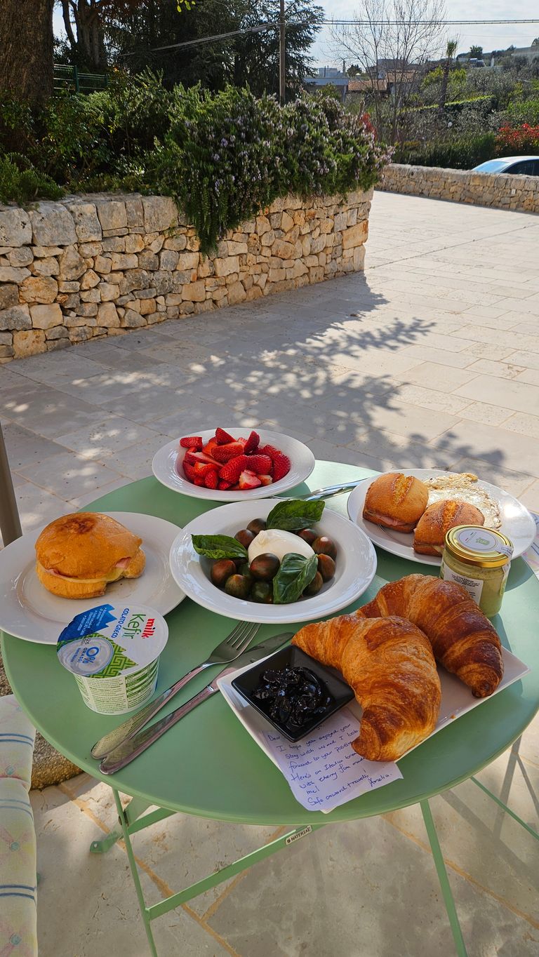 An outdoor table set with various foods including croissants, a sandwich, a bowl of strawberries, a plate with tomatoes and buffalo mozzarella, a yogurt cup, jam, and a jar of honey in front of a stone wall and summer garden.