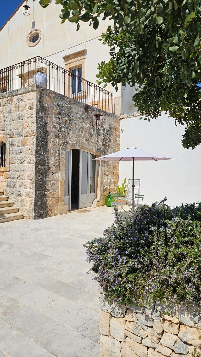 Sunny patio with stone wall, flower box with purple blossoms, white sun umbrella, and metal table with chairs in front of a building with a light plastered upper floor and stone lower section.
