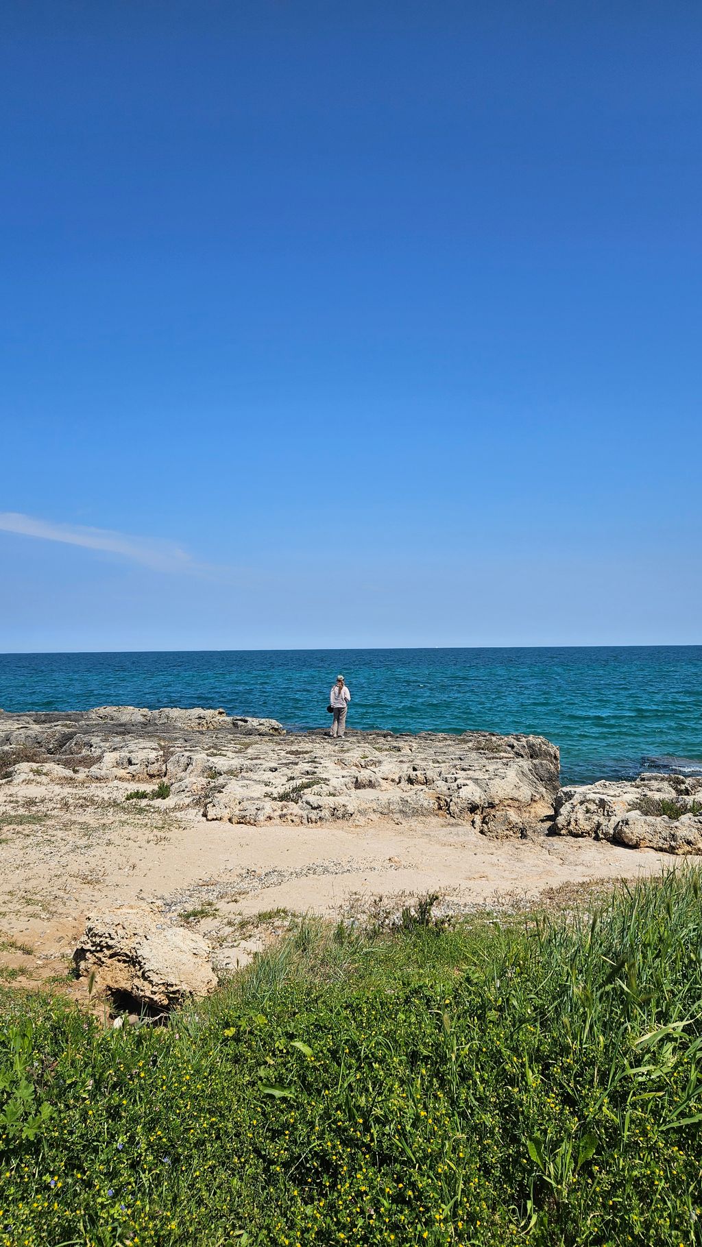 A lone person stands on rocky shore in front of the clear blue sea on a sunny day with blue sky.
