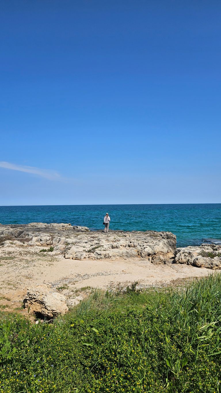 A lone person stands on rocky shore in front of the clear blue sea on a sunny day with blue sky.
