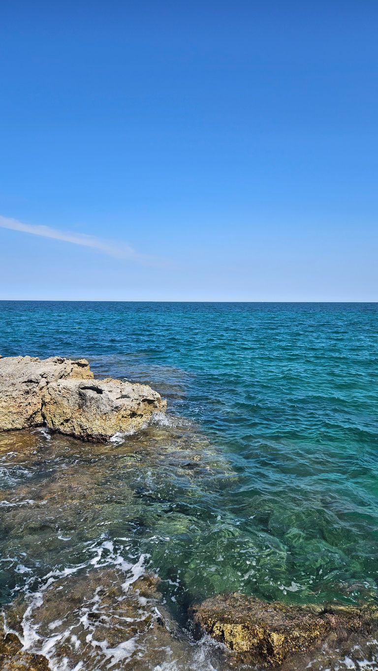 Clear blue ocean with rocks in the foreground and a cloudless sky.