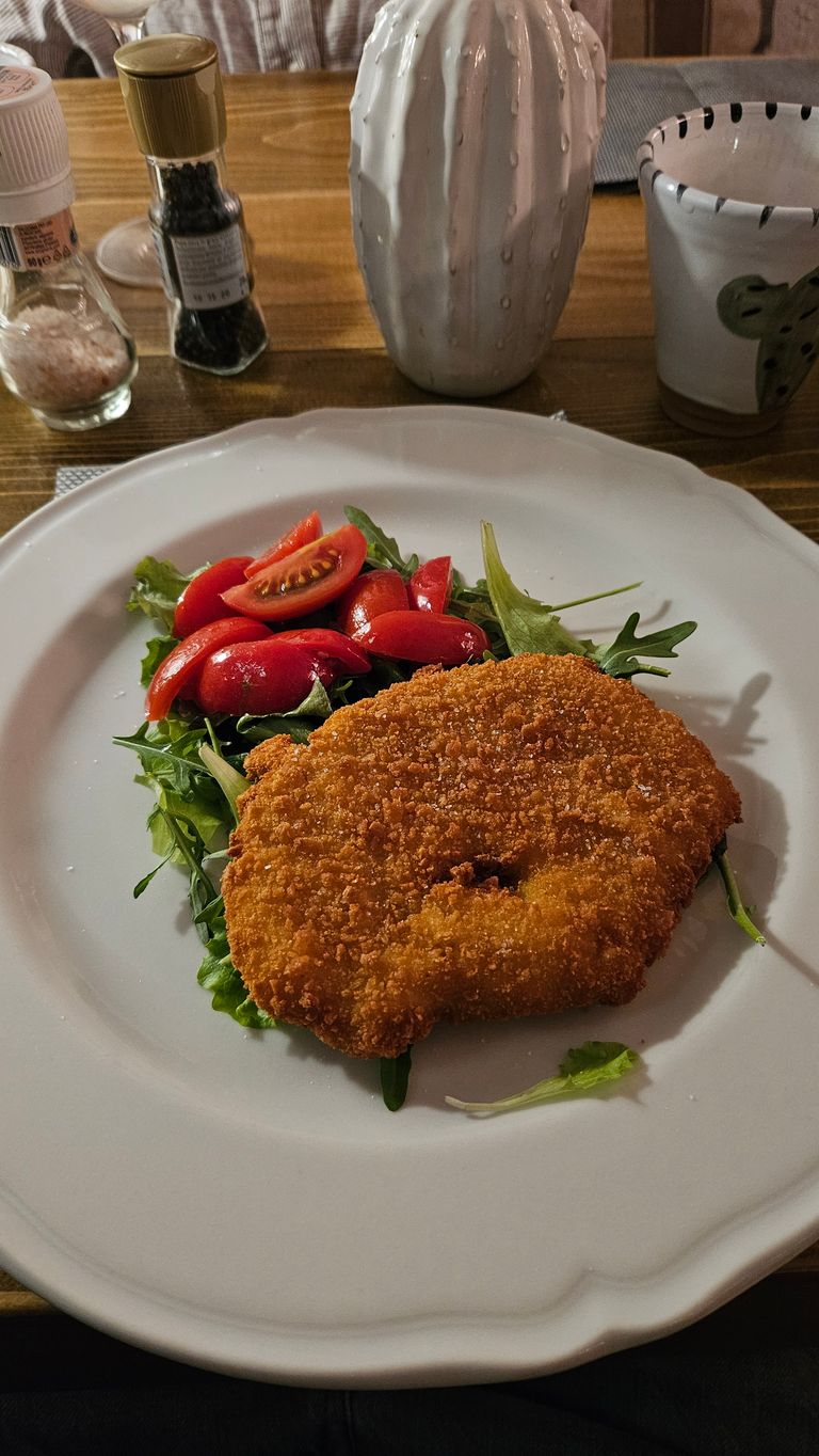 Breaded schnitzel served with a small salad of arugula and halved cherry tomatoes on a white plate on a wooden table.