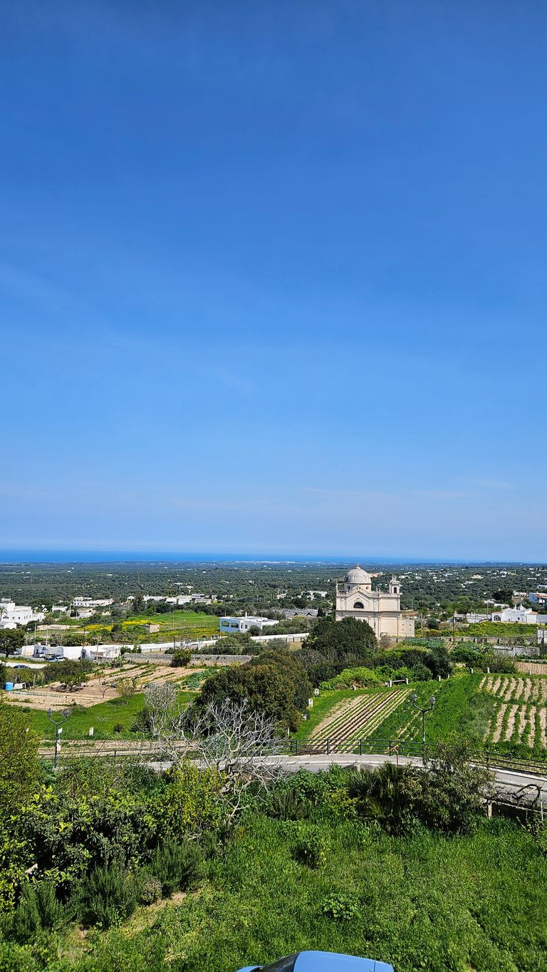 Landscape view of a rural area with fields, a small historic building with a dome, and a distant view of the sea under a clear blue sky.