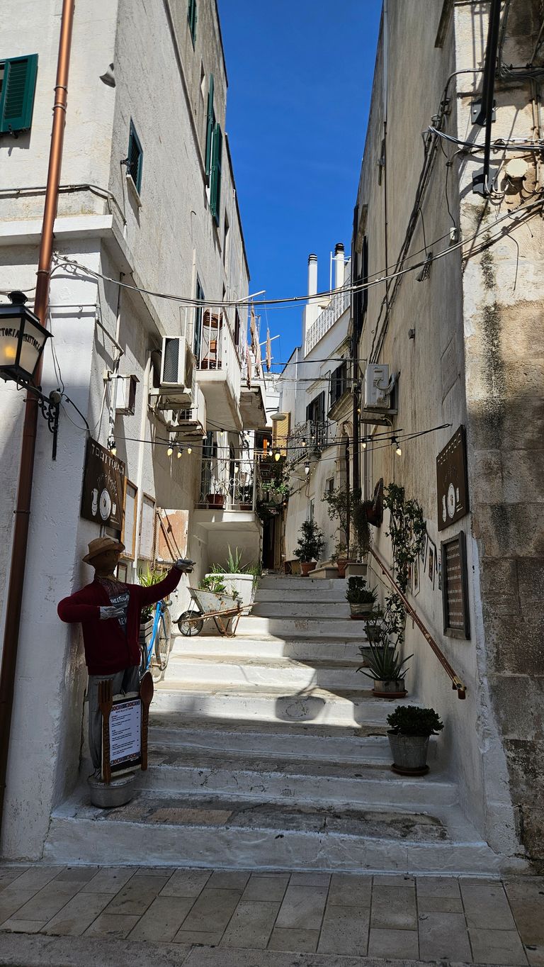 Narrow stairway street between white buildings with several potted plants and a figure in the left corner holding a sign.