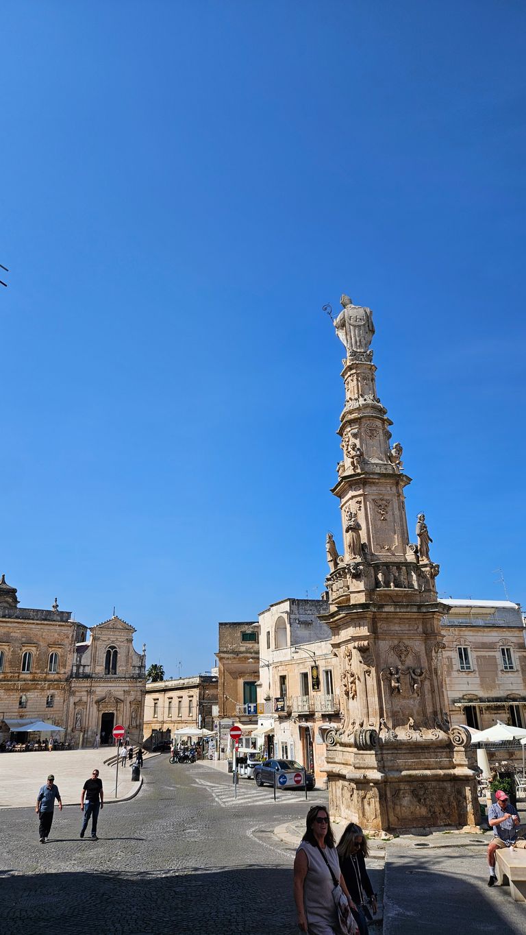 A tall, ornate stone column obelisk on a square with historic buildings in the background and several people walking around it under a clear blue sky.