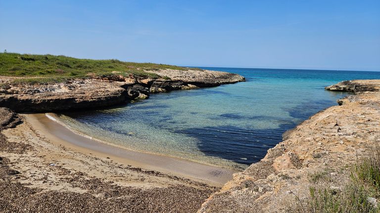 Small beach with clear water surrounded by rocky coastline and green grass under a blue sky.