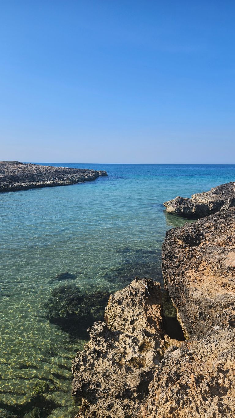 Clear turquoise sea water next to rugged, brown rocks under a cloudless blue sky.
