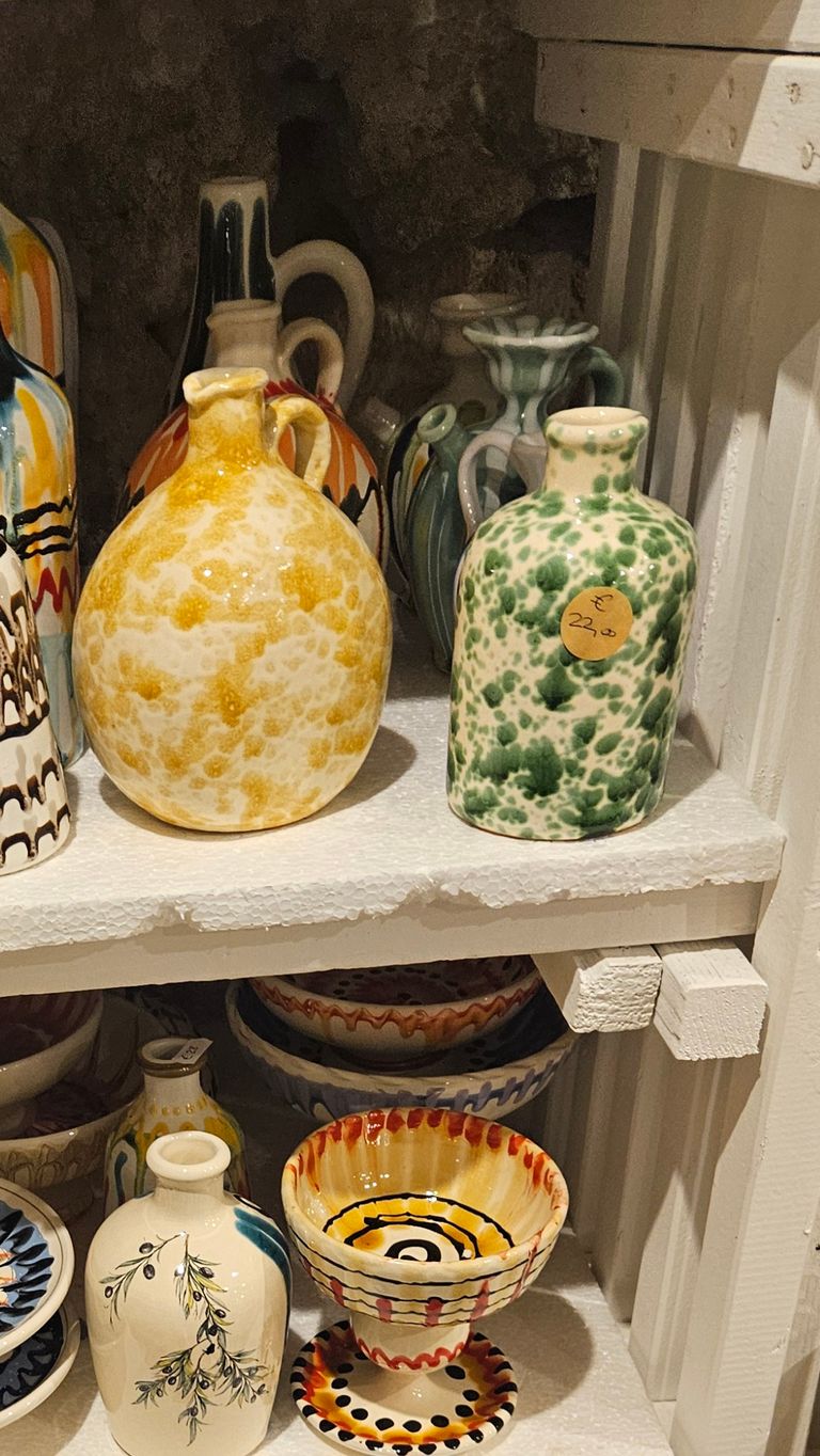 Several colorful ceramic bottles and bowls displayed on white wooden shelves, including two prominent bottles with yellow-white and green-white speckled patterns.