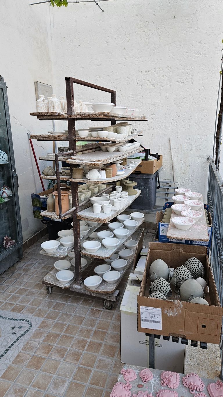 Multi-tiered shelf holding white ceramic bowls and pots in a workshop by a wall, with cardboard boxes containing clay objects and decorated pink ceramic pieces on the floor.