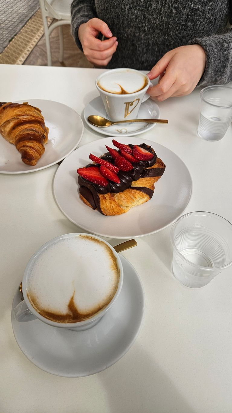 Coffee setting with two cups of cappuccino, a croissant, and a chocolate pastry topped with sliced strawberries on a white table, a person wearing a grey sweater holding one cup.