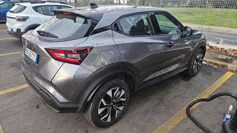 A silver-gray compact Nissan Qashqai SUV parked in a parking lot, viewed from the rear right side showing the taillights, rear wheel, and license plate.