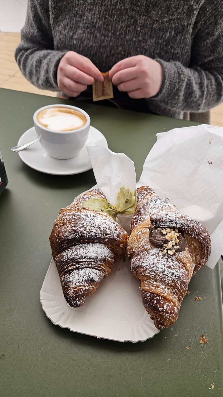 Two powdered sugar-coated croissants served on brown paper, next to a cup of cappuccino on a white plate and a person in a gray sweater opening a sugar packet.