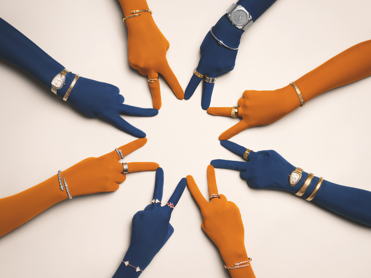 Hände in blauen und orangen Handschuhen mit Ringen, Armreifen und Armbanduhren aus Gold, Edelstahl und Diamanten.