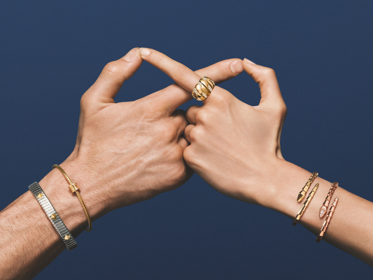 Zwei Hände vor dunkelblauem Hintergrund: goldener gewölbter Ring am Mittelfinger, goldenes Gliederarmband und silberfarbenes Band mit Goldakzenten am Handgelenk, rosegoldene Schlangenarmreifen am Arm.
