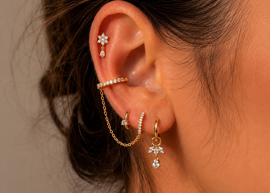 Ohr mit goldenen Ohrschmuckstücken: Blütenstecker am Helix, Zirkonia-Ear-Cuff mit Kettenverbindung, Creolen und Anhänger mit weißen Steinen.