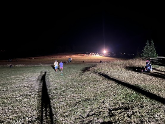Campingwiese in der Nacht. Langer Schlagschatten einer Person unter einem Flutlicht. Zwei Personen kommen entgegen. Weit im Hintergrund ein Lichtkegel eines anderen Flutlichts. Dazwischen und daneben: Dunkelheit. 