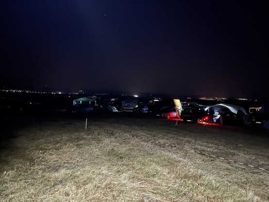 Braun grüne Wiese von einem Lichtkegel erhellt. In der Mitte Campingfahrzeuge und Zelt mit diffusen bunten Lichtquellen. Im Hintergrund beleuchtet Ortschaften 
