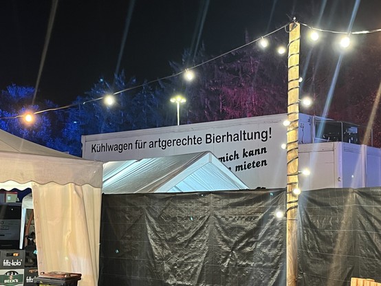 Freiluft Gästebereich. Lichterkette an Holzmasten, ein weiser Pavillon, ein Zaun mit schwarzer Sichtschutzplane im Hintergrund ein weißer Kühlwagen mit der Aufschrift "Kühlwagen für artgerechte Bierhaltung"