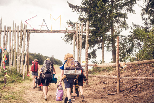 Personen gehen mit Gepäck beladen einen Feldweg entlang durch ein Holztor über dem steht "Camp"