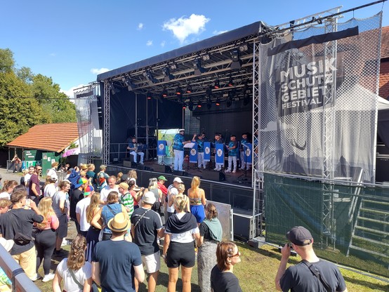 Festivalbesucher vor einer Bühne auf der ein Shanty Chor singt. Die Chormitglieder tragen einheitlich Schifffahrtsmützen, dunkelblaue Hemden mit Segelschiff und weise Hosen.