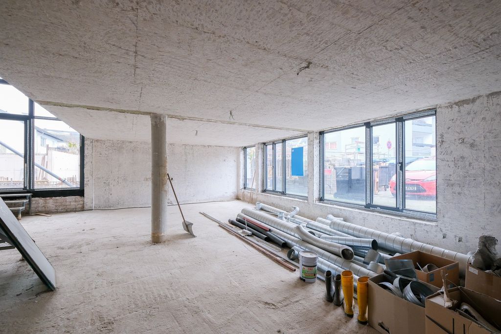 Empty unfinished room with concrete walls and ceiling, large windows, a central support column, and pipes and construction materials scattered on the floor.