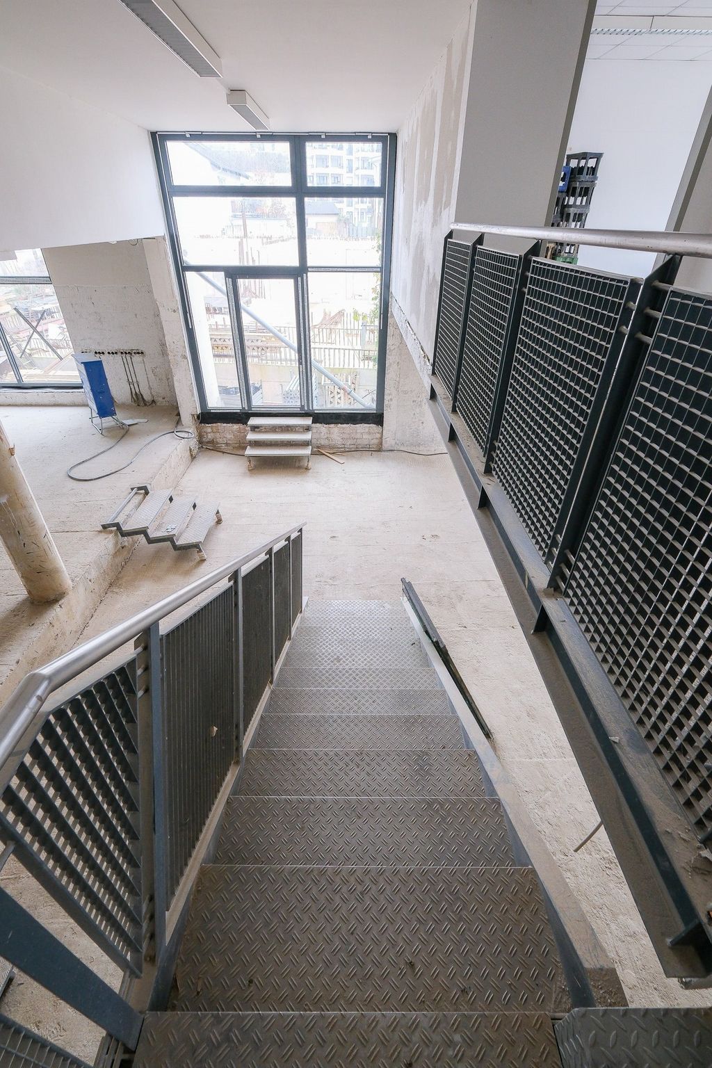 View from above of a metal staircase with grid railings in an unfinished or renovation-needed room with large windows.