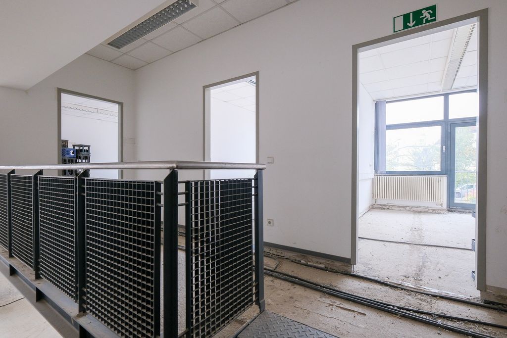 Hallway in an unfurnished office building with gray metal railings, white walls, two doorways, and large windows showing trees outside.