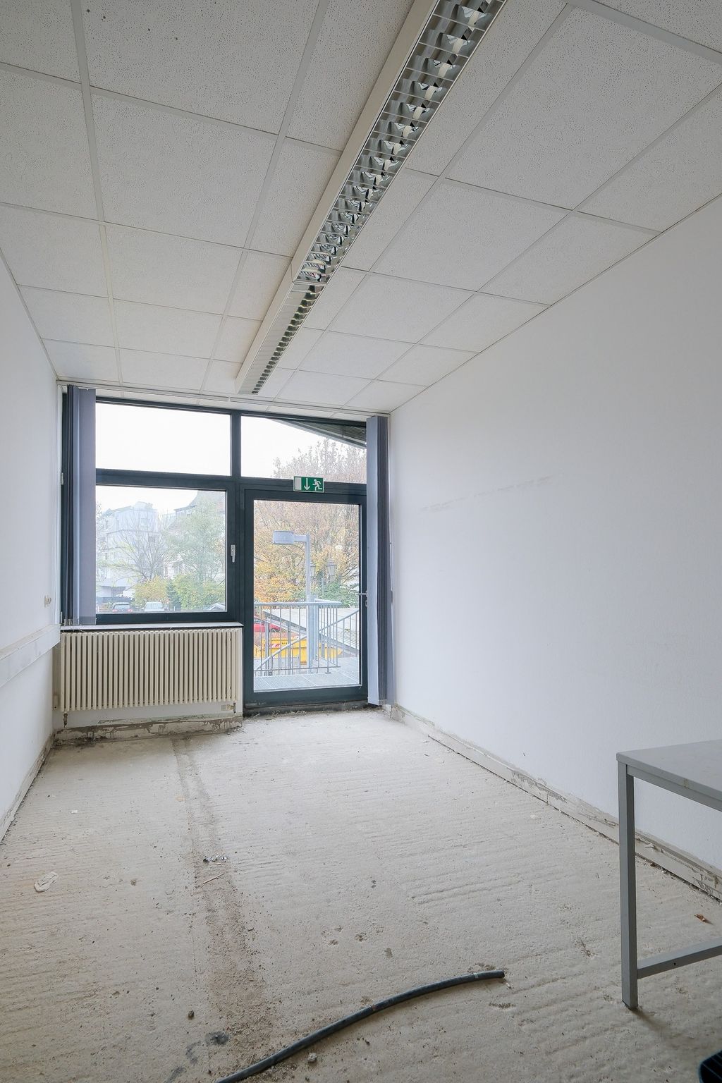 Empty unfinished room with bare concrete floor, white walls, a large window, and a glass door with a green emergency exit sign showing an outdoor staircase and trees outside.