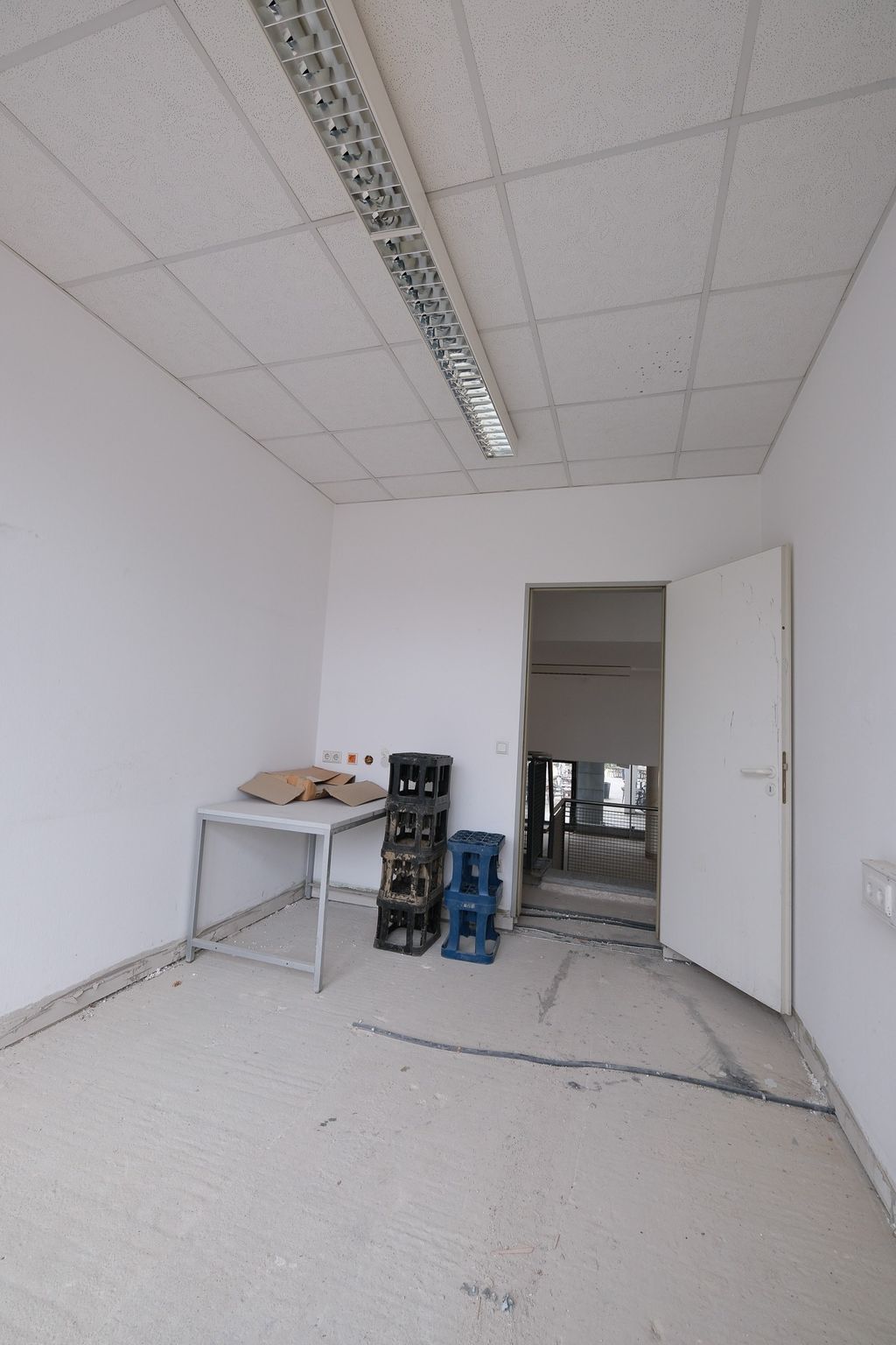 Empty unfinished room with a white table, three stacked plastic crates, and an open door leading to a stairwell.