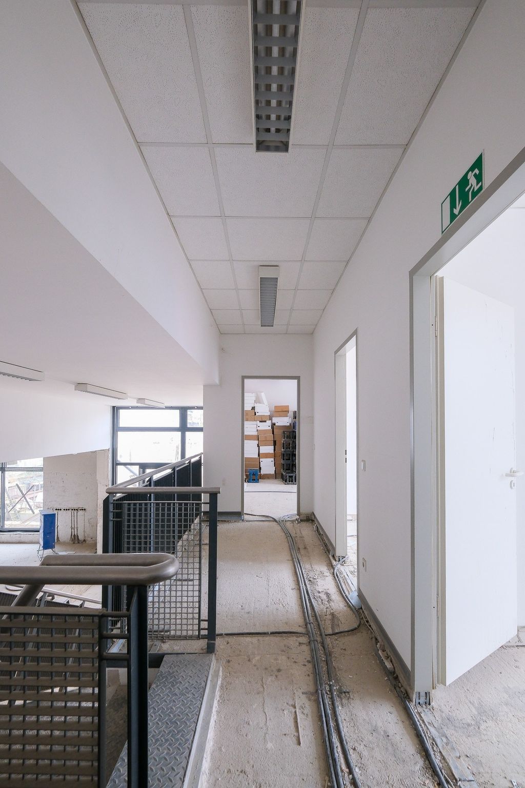 Interior view of a partially finished hallway with exposed cables on the floor, an open stair railing, and several doors, one leading to a room stacked with boxes.