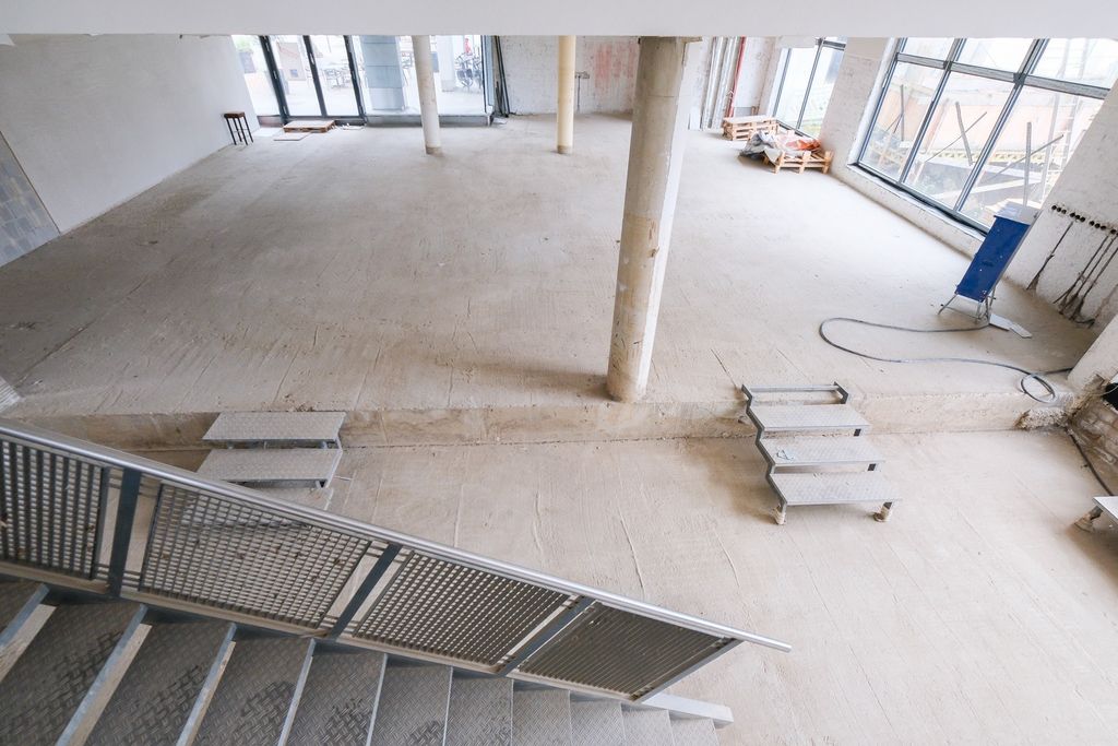 Interior of an unfinished construction site with raw floor, stairs, and several columns, large window in the background showing scaffolding outside.