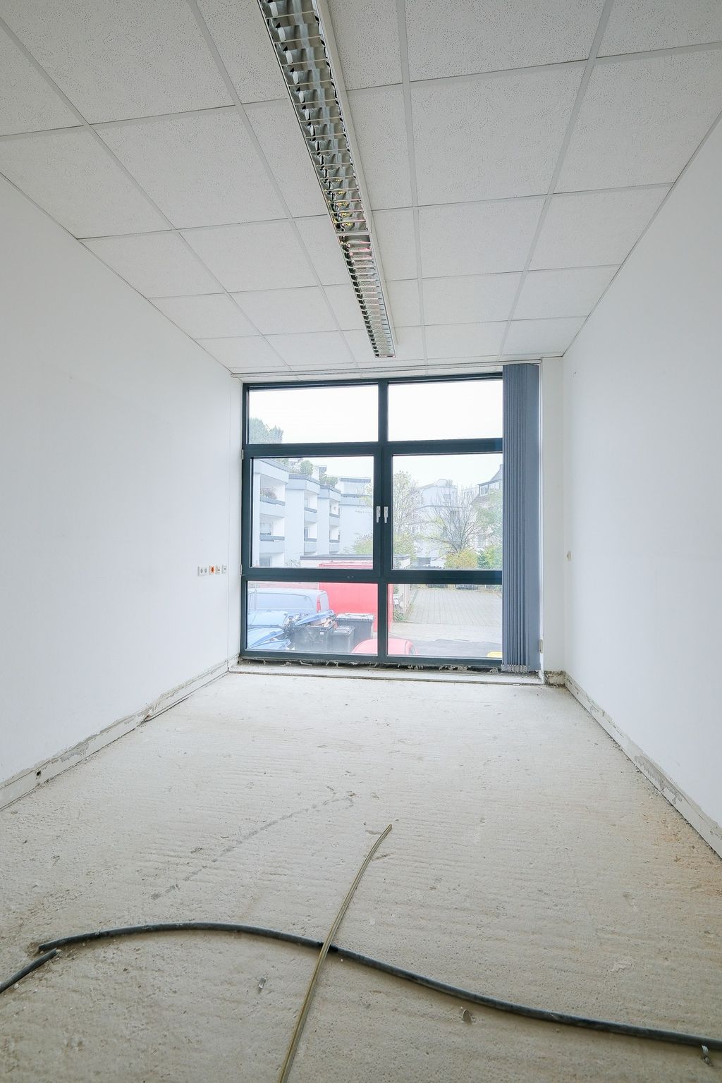 Empty, unfinished room with white walls and a large window showing parked vehicles and buildings outside.