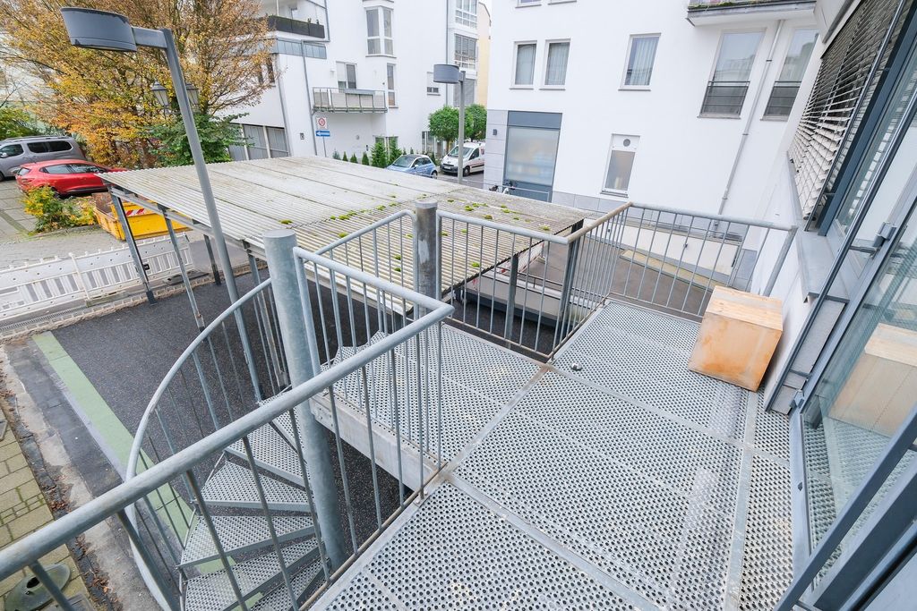 Elevated rectangular metal terrace with perforated flooring and surrounding railing, a wooden block in the corner, a spiral staircase leading down, surrounded by residential buildings and parking spaces with autumn trees in the background.