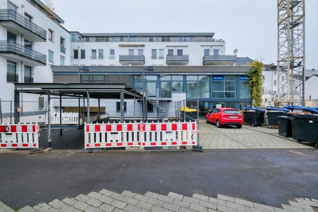 Modern residential buildings with light facades and balconies in the background, in front a construction site with barriers and a covered area, to the right a parking lot with garbage bins and a red car.