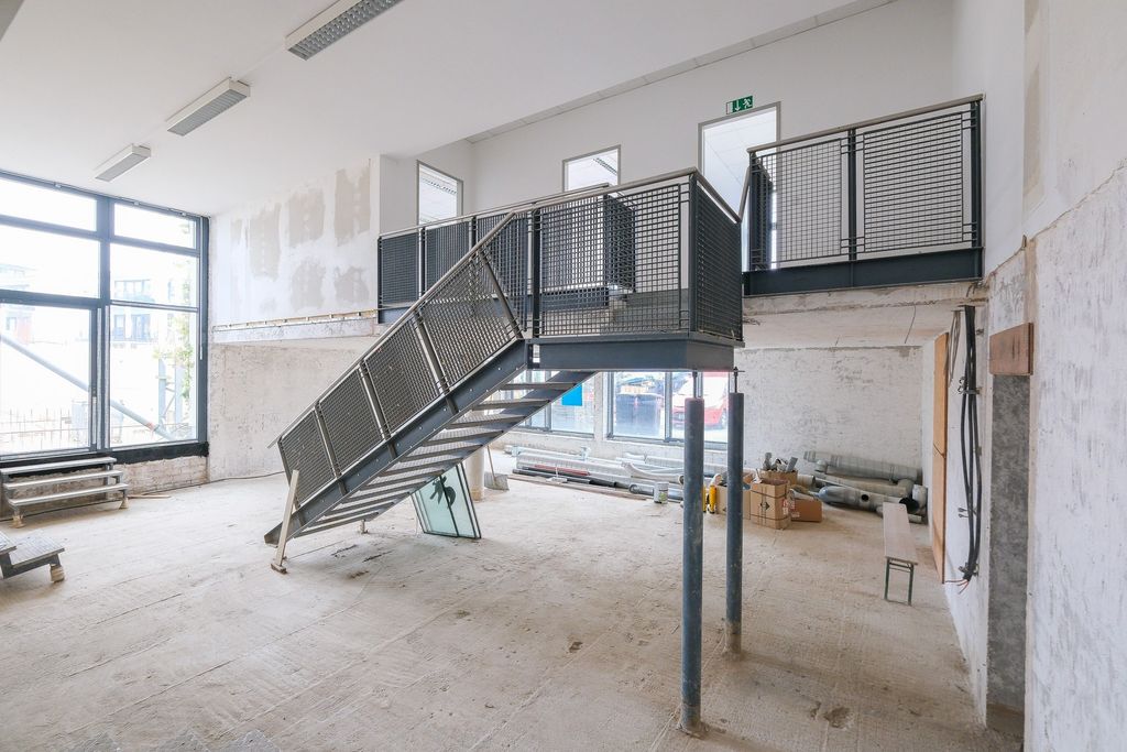 Interior of an empty unfinished building with a metal staircase leading to a mezzanine. Large windows let in daylight, the floor is raw and walls are partially plastered.