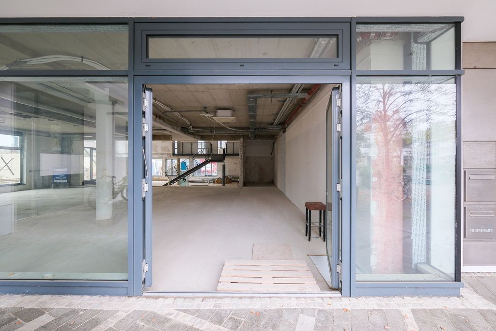 Empty modern commercial space with large glass windows and blue door frames, visible ceiling pipes, a small wooden pallet and a stool inside.