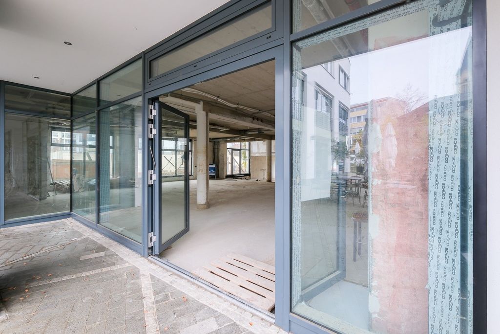 A large empty room with concrete floors and columns behind a glass facade with an open glass door, located on the ground floor of a building.