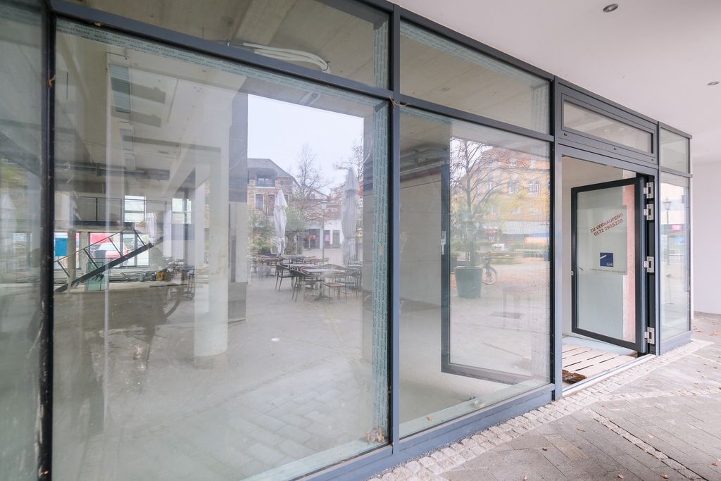 Reflective glass windows of a modern building overlooking an empty courtyard with tables, chairs, and closed umbrellas on a cloudy day.