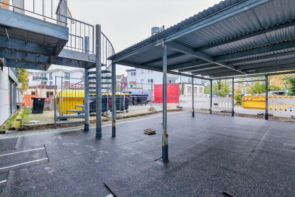 Covered outdoor area with a steel structure and spiral staircase in an urban setting with waste containers and construction site elements in the background.