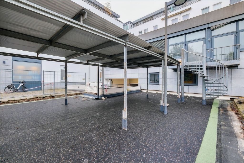 Outdoor area with a large covered section supported by metal poles, a bicycle parked near buildings, and a spiral outdoor staircase attached to the building wall.