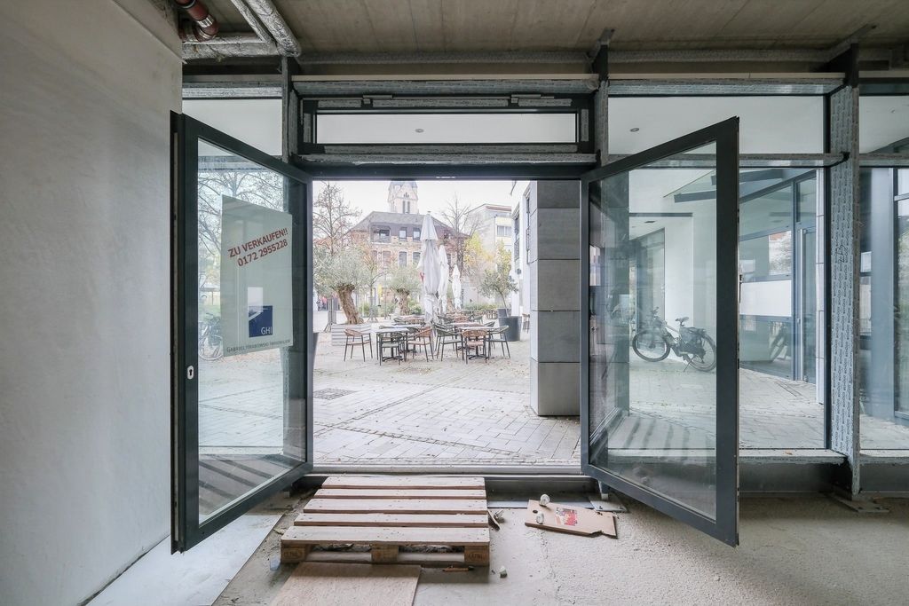 View through an open glass door onto a paved courtyard with chairs, tables, and a closed parasol, with a wooden pallet and a box with paper on the floor inside.